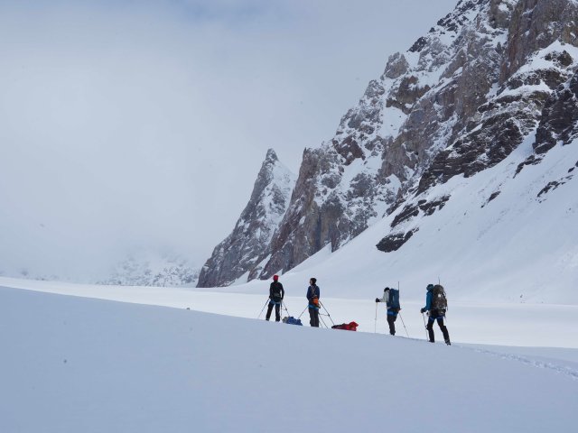 "Four skiers dragging their field equipment up a snowy hill, next to a mountain"